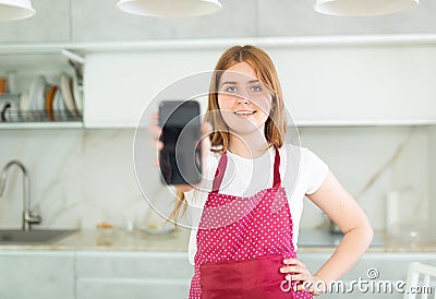 in kitchen,smiling red-haired girl in polka-dot apron shows blank smartphone screen close-up Stock Photo