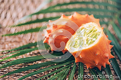 Kiwano or african horned melon with palm leaves on rattan background. Cutted hedged gourd, african horned cucumber, english tomato Stock Photo