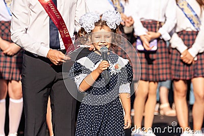 First-form schoolgirl at school on holiday of beginning of elementary education reading poetry. Girl with microphone declaiming Editorial Stock Photo