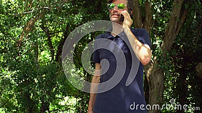 L'homme mûr émotif attirant avec la barbe et les lunettes de soleil parle par le smartphone dans la forêt ou le parc clips vidéos L'homme mûr émotif attirant avec la barbe et les lunettes de soleil parle par le smartphone dans la forêt ou le parc clips vidéos