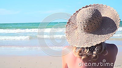 La mujer rubia joven con el sombrero azul del bikini y de paja se sienta en la playa y mira el mar almacen de metraje de vídeo La mujer rubia joven con el sombrero azul del bikini y de paja se sienta en la playa y mira el mar almacen de metraje de vídeo