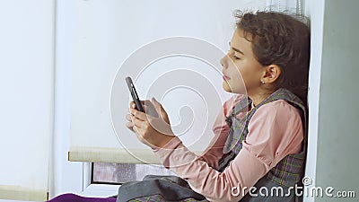 La ragazza teenager che si siede sul web un davanzale della finestra gioca il gioco online per lo smartphone video d archivio La ragazza teenager che si siede sul web un davanzale della finestra gioca il gioco online per lo smartphone video d archivio