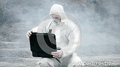 A laboratory technician in a mask and a chemical protective suit opens a toolbox on dry land, around toxic smoke Stock Photo