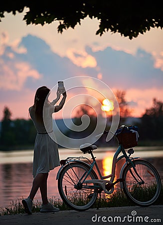 Lady near retro bike takes sunset photo on cell phone near the lake Stock Photo