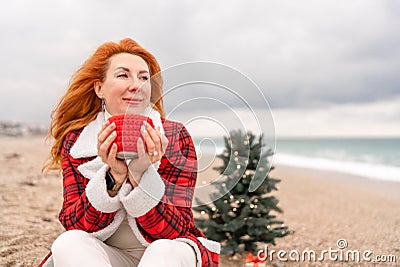 Lady in plaid shirt with a red mug in her hands enjoys beach with Christmas tree. Coastal area. Christmas, New Year Stock Photo