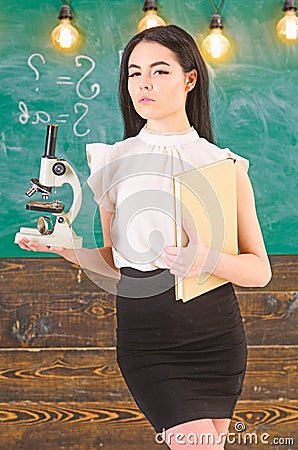 Lady scientist holds book and microscope, chalkboard on background, copy space. Lady in formal wear on calm face in Stock Photo
