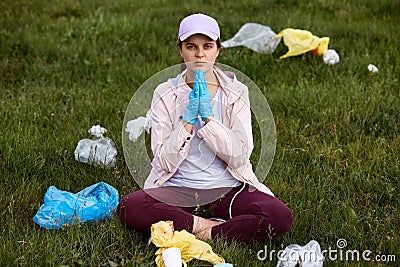 Lady sitting on ground in green meadow and praying for better for environment, keeping hands together surrounded with trash, being Stock Photo