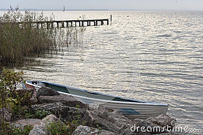 Lake shore with boat moored between large boulders. Background with water, wooden jetty and background sailboat. Stock Photo