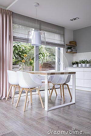 Lamp above wooden table and white chairs in dining room interior with drapes and window. Real photo Stock Photo