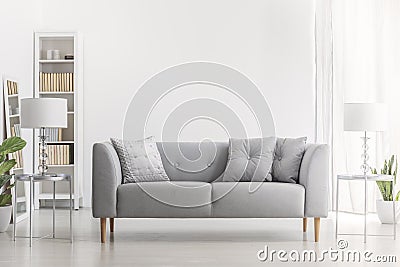 Lamp on silver table next to grey sofa with pillows in white living room interior with plant. Real photo Stock Photo