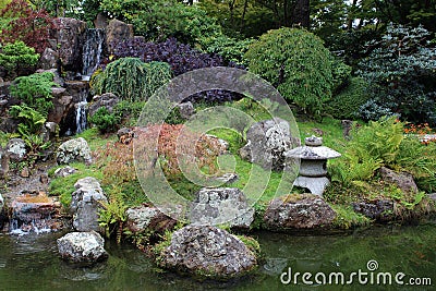 A landscaped Japanese garden with a waterfall and various trees, shrubs, grasses, ferns and a stone lantern Editorial Stock Photo