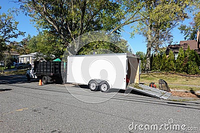 A landscaping truck with a long white enclosed trailer trailer with it`s rear ramp down seen on a shady residential asphalt stree Stock Photo