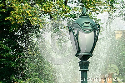 Lantern or park light against water jet spray from fountain and green trees. Modern city street landscape in sunny summer day Stock Photo