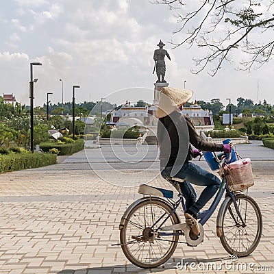 A Laotian woman rides a bicycle in the Chao Anouvong park with the large statue of King Chao Anouvong in the background in Vientia Editorial Stock Photo