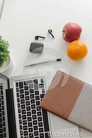 Laptop, black wireless earbuds, notebook, pen, potted plant, apple and orange on white background Editorial Stock Photo