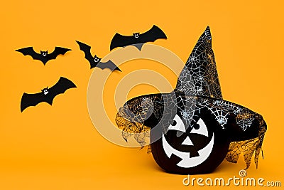 Large Black Halloween Pumpkin with cute smiling face wearing witch hat looking at flying bats over orange background. Stock Photo