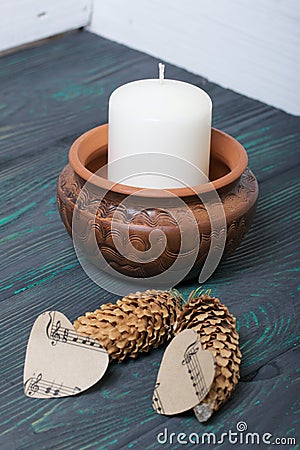 A large candle in a clay vessel. Decorated with cones and hearts cut out of paper. It stands on painted boards painted in black Stock Photo