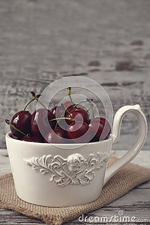 A large cup of coffee in front angel, white bowl full with fresh cherries, fruits. Light rustic background, shabby chic, vintage Stock Photo