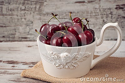 A large cup of coffee in front angel, white bowl full with fresh cherries, fruits. Light rustic background, shabby chic, vintage Stock Photo