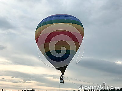 Large multi-colored bright round rainbow colored striped striped flying balloon with a basket against the sky in the evening Stock Photo