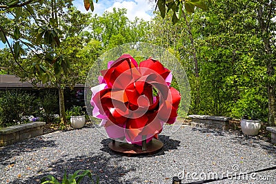A large red and pink metal flower in the garden surrounded by lush green trees and plants with blue sky and clouds Editorial Stock Photo