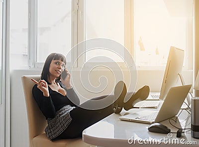 HOT brunete talking at the phone in office outfit sitting at the table with a computer and a laptop on it Stock Photo