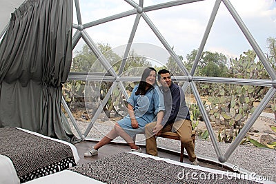 Latin couple in love inside glamping dome room with desert vegetation outside Stock Photo