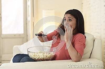 Latin woman sitting at home sofa couch in living room watching television scary horror movie Stock Photo