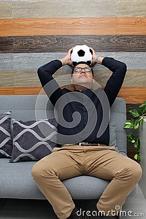 Latino adult man with glasses watches soccer game on TV on his sofa with his ball and gets excited, angry, sad watching the soccer Stock Photo