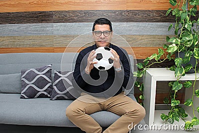 Latino adult man with glasses watches soccer game on TV on his sofa with his ball and gets excited, angry, sad watching the soccer Stock Photo