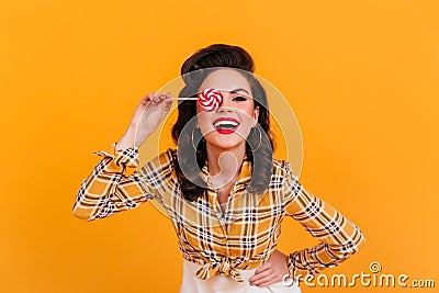 Laughing curly woman posing with candy. Studio shot of excited pinup lady with lollipop on yellow background Stock Photo