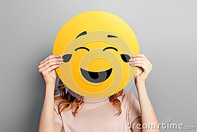 Laughing emoticon with tears of joy. Girl holds a yellow smiley with romantic love face emoticon on a grey Stock Photo