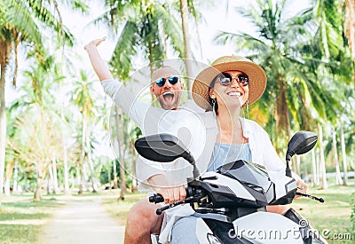 Laughing happy couple travelers riding motorbike during their tropical vacation under palm trees. Man emotionally raised hand up Stock Photo