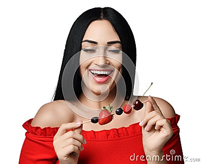 Happy smiling brunette woman in red off shoulder dress holding a small skewers with berries isolated on white. Stock Photo