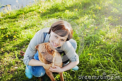 Laughing happy young woman in denim overalls hugging her red cute dog Shar Pei in the green grass in sunny day, true friends Stock Photo