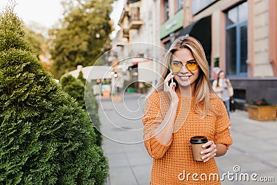 Laughing pretty lady talking on phone while walking by bushes with cup of tea. Outdoor portrait of ecstatic blonde woman Stock Photo