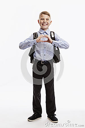 Laughing schoolboy in uniform with a backpack shows his heart with his hands. Back to school. White background. Vertical Stock Photo