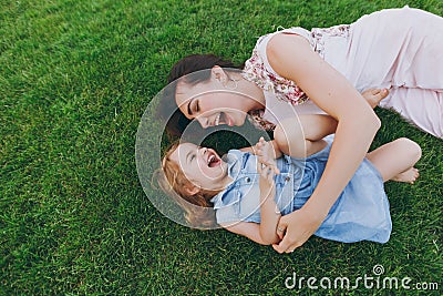 Laughing woman in light dress and little cute child baby girl lie on green grass lawn tickle, play and have fun. Mother Stock Photo