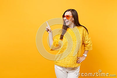 Laughing young woman in fur sweater, heart orange glasses pointing index finger aside on copy space isolated on bright Stock Photo
