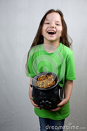 Laughting cute seven years old girl with green shamrok leaf on her cheek showing off cast iron pot, full of leprechaun gold coins Stock Photo