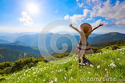 On the lawn in mountains landscapes the hipster girl in dress, stockings and straw hat stays watching the sky with clouds. Stock Photo