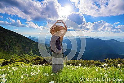 On the lawn of white flowers the hipster girl is staying in long dress, straw hat with back sack, enjoying the sunset. Stock Photo