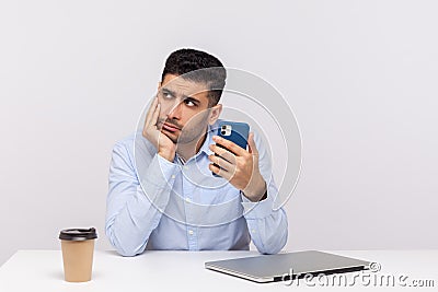 Lazy inefficient unproductive man employee sitting in office workplace, holding smartphone and looking away Stock Photo