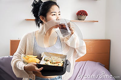 Lazy Asian woman eating junk food hand holding box of pizza and bowl of potato chips drinking sweet soda in bed Stock Photo