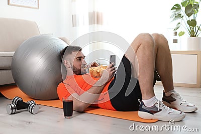 Lazy young man with smartphone eating junk food on yoga mat Stock Photo