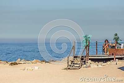 View of technological senior woman taking sunbath and using her mobile phone on beach club, sea as background, in Portugal Editorial Stock Photo