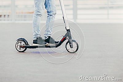 Legs of unknown male in black sneakers and jeans rides on electric scooter over urban asphalt, enjoys sunny day. People and leisur Stock Photo