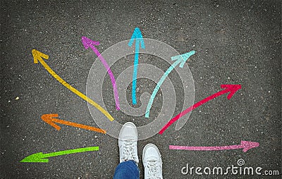 Legs in white sneakers in front of colorful arrows pointing in different directions on the gray asphalt Stock Photo