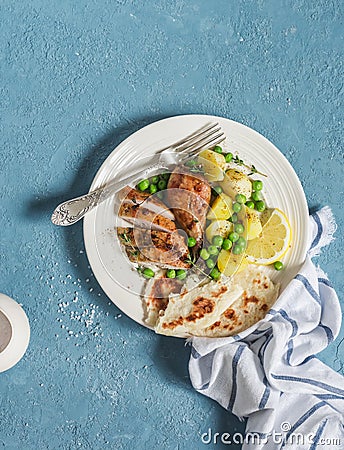 Lemon thyme baked chicken, potatoes and green peas on a white plate on a blue background. Stock Photo