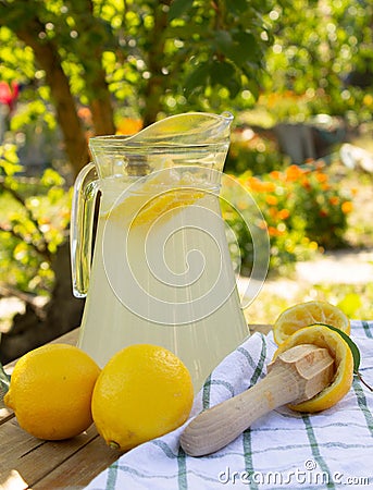 Lemonade in glass jug / pitcher on table in the garden on a sunny day. Ripe lemons, wooden citrus pestle and towel on wooden table Stock Photo